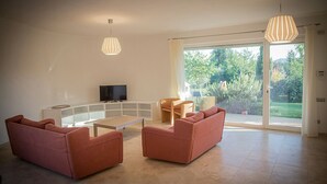 A modern living room with a glass coffee table, two sofas, a TV, and a large sliding glass door leading to a garden.