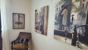 Une pièce comprenant une chaise en bois, un tableau représentant un paysage urbain accroché au mur, et un autre tableau représentant un couple tenant un verre de vin.