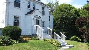 Une maison blanche à deux étages, dotée d’un porche à l’avant et d’un escalier menant à l’entrée.