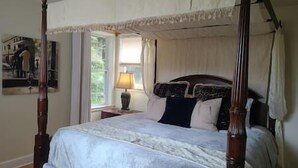 Un lit à baldaquin avec un couvre-lit blanc et une tête de lit sombre, une table de chevet avec une lampe et une fenêtre avec des rideaux.