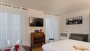 A hotel room with a bed, a TV mounted on the wall, a wooden dresser, a desk with a lamp, and a cityscape wall art. - Superba Studio by Wonderful Italy (Genoa)