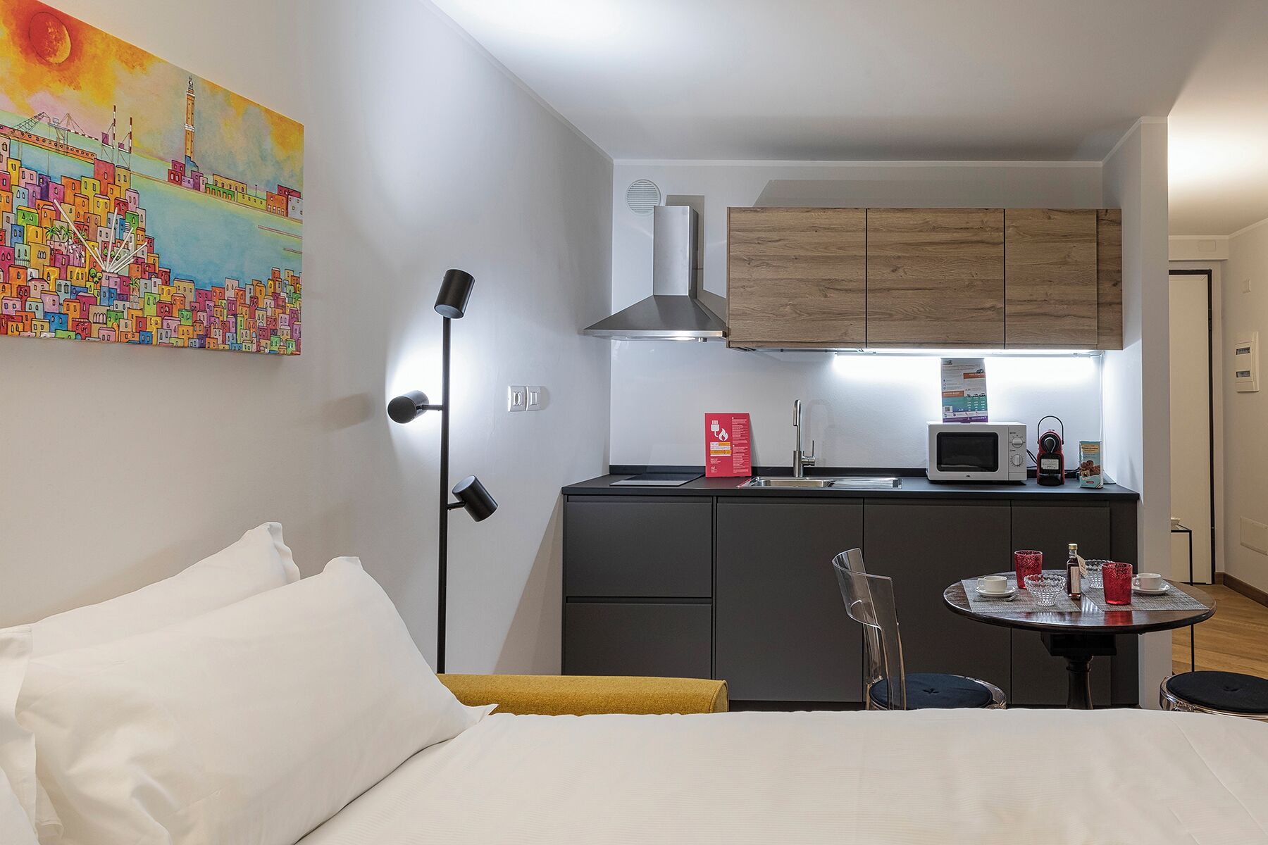 A compact hotel room with a bed, a small kitchenette, a dining table with chairs, and a colorful cityscape painting on the wall.