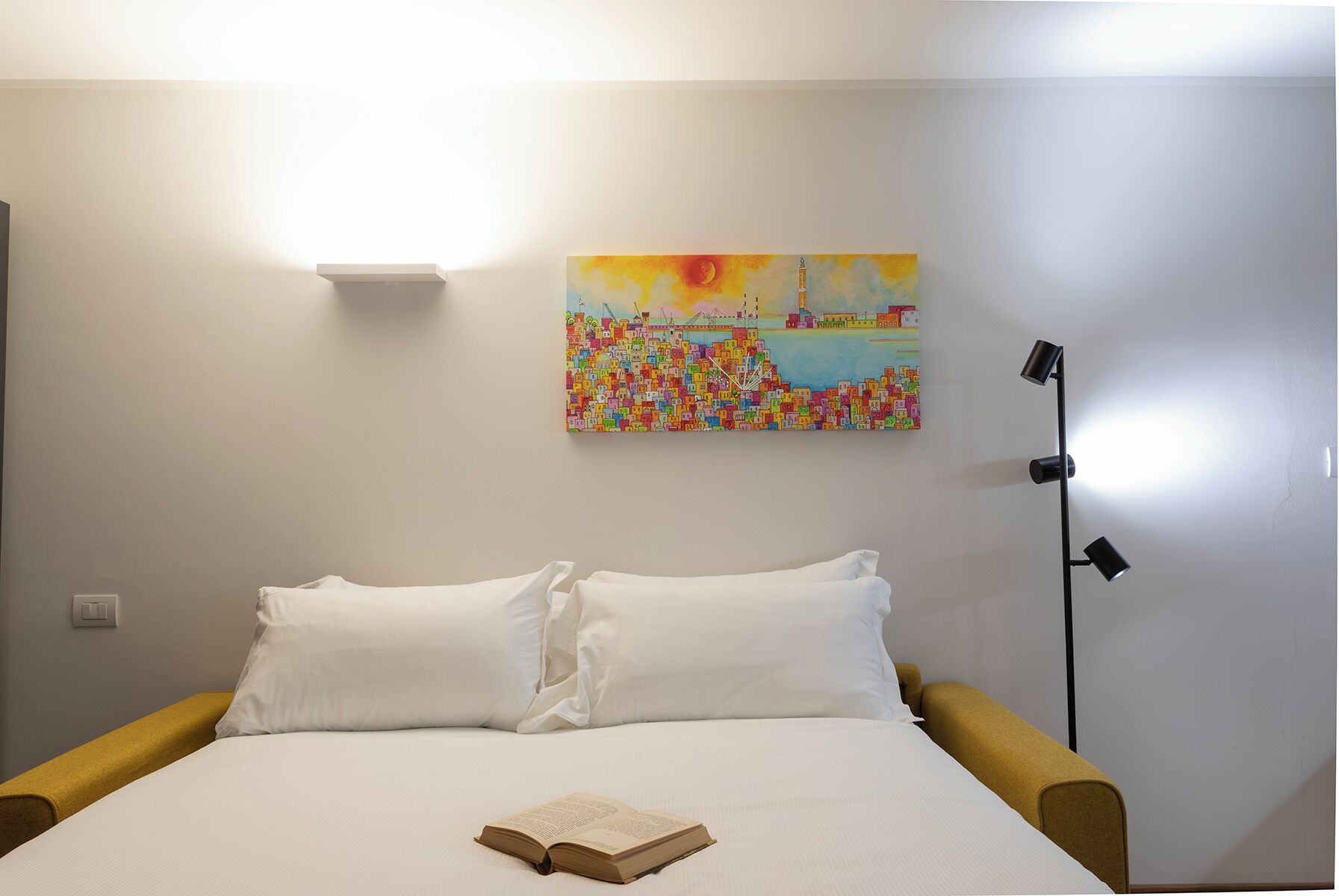 A hotel room with a bed, pillows, a book, a painting, and a floor lamp.