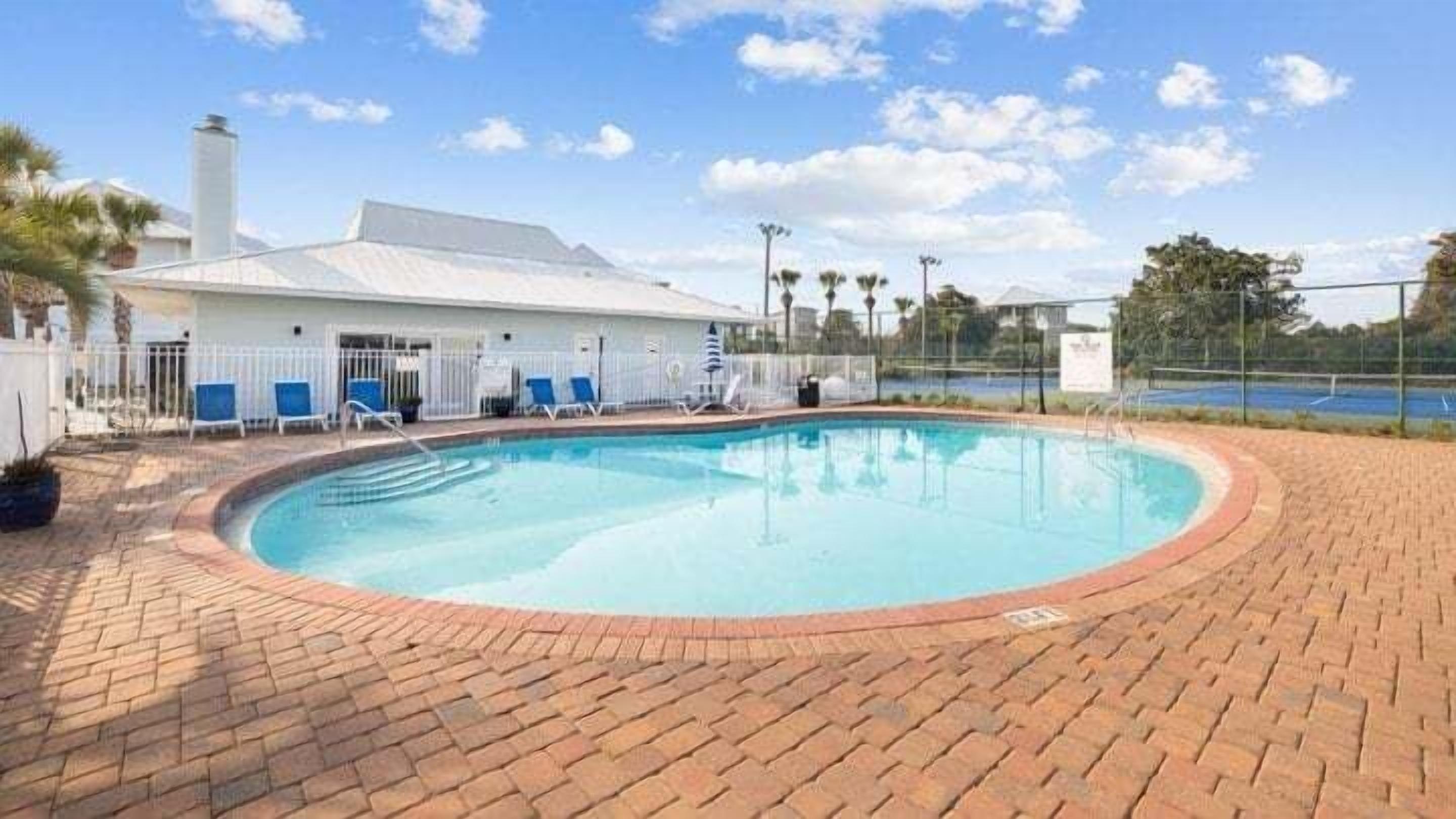 A swimming pool with a brick border, a white building with a flat roof, and blue chairs.