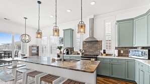 A modern kitchen with a large island, bar stools, hanging pendant lights, and a view of the city. - Oasis Vacations, Sweet Tater, Pool, Elevator, Putting Green, Marsh Views (Ocean Isle Beach)