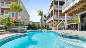 A swimming pool surrounded by multi-story buildings with balconies, stone planters with flowers, and outdoor seating areas. - Oasis Vacations, Sweet Tater, Pool, Elevator, Putting Green, Marsh Views (Ocean Isle Beach)