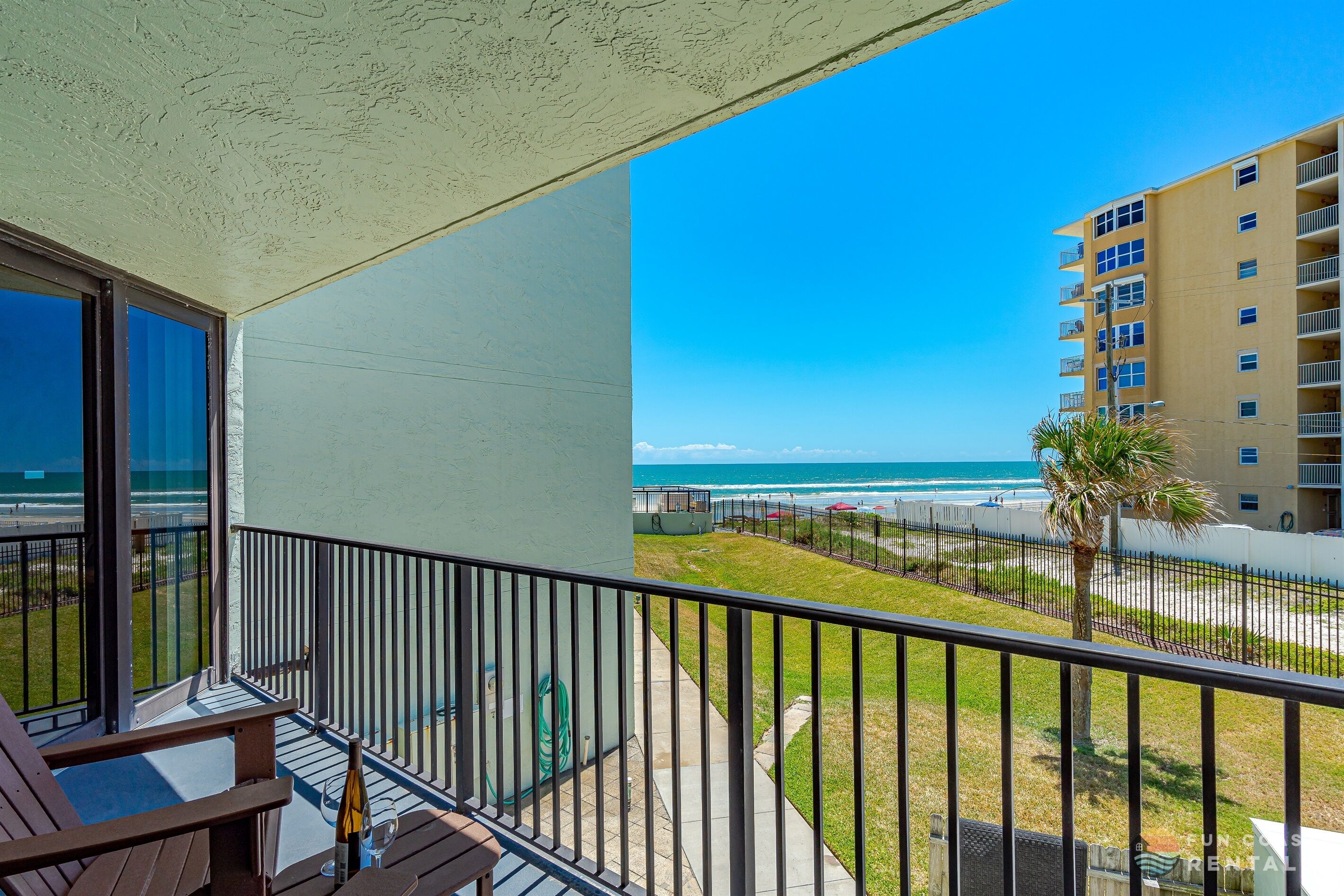A balcony with a view of the beach and ocean, a palm tree, and a building in the background.