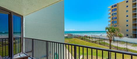 A balcony with a view of the beach and ocean, a palm tree, and a building in the background.