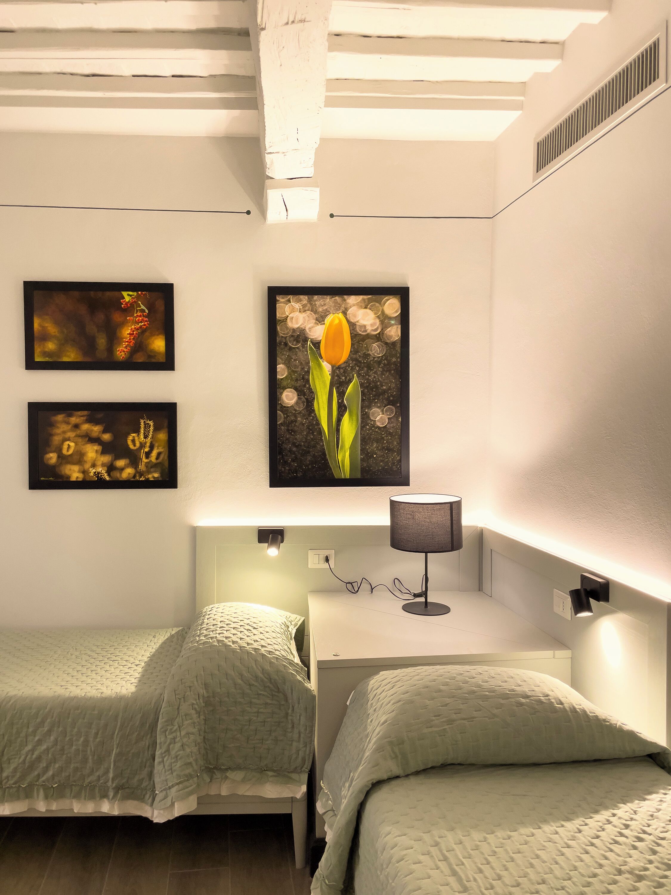 A bedroom with two beds, a nightstand with a lamp, and three framed artworks on the wall.
