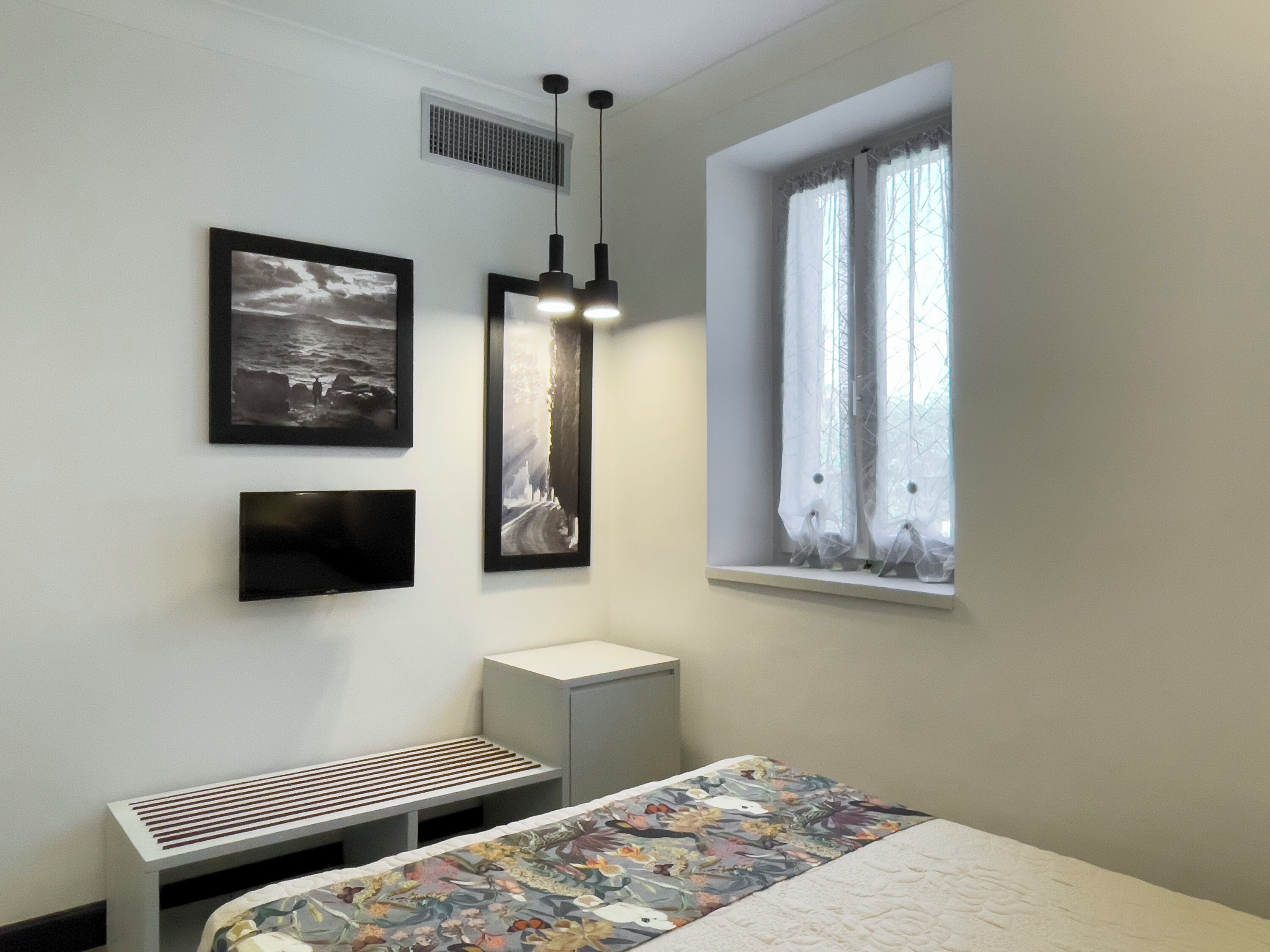 Quarto com cama, um banquinho, janela com cortinas e duas luzes fixadas na parede.