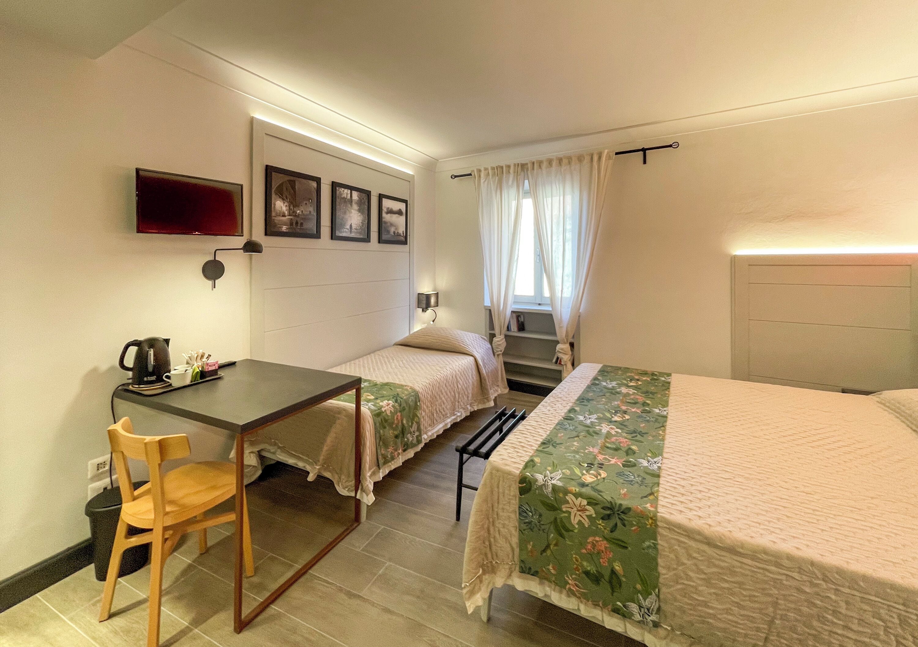 A small, well-lit room with two single beds, a desk with a chair, a TV mounted on the wall, and a window with curtains.