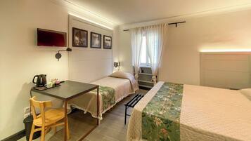 A small, well-lit room with two single beds, a desk with a chair, a TV mounted on the wall, and a window with curtains.