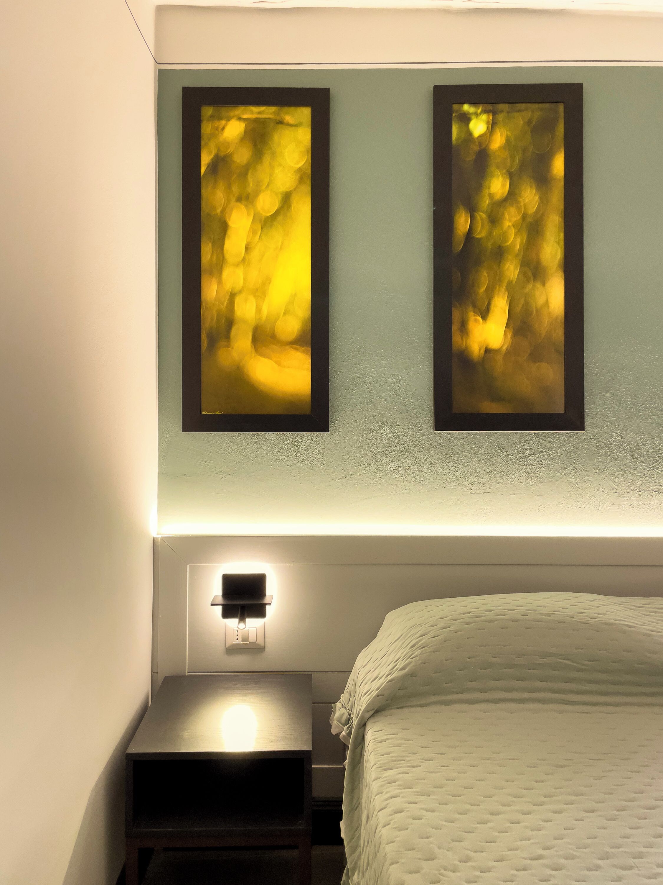 A hotel room with a bed, a bedside table with a lamp, and two framed abstract paintings on the wall.
