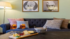 Una sala de estar con un sofá azul oscuro, dos cuadros enmarcados, una lámpara, una mesa con queso, uvas y bebidas.