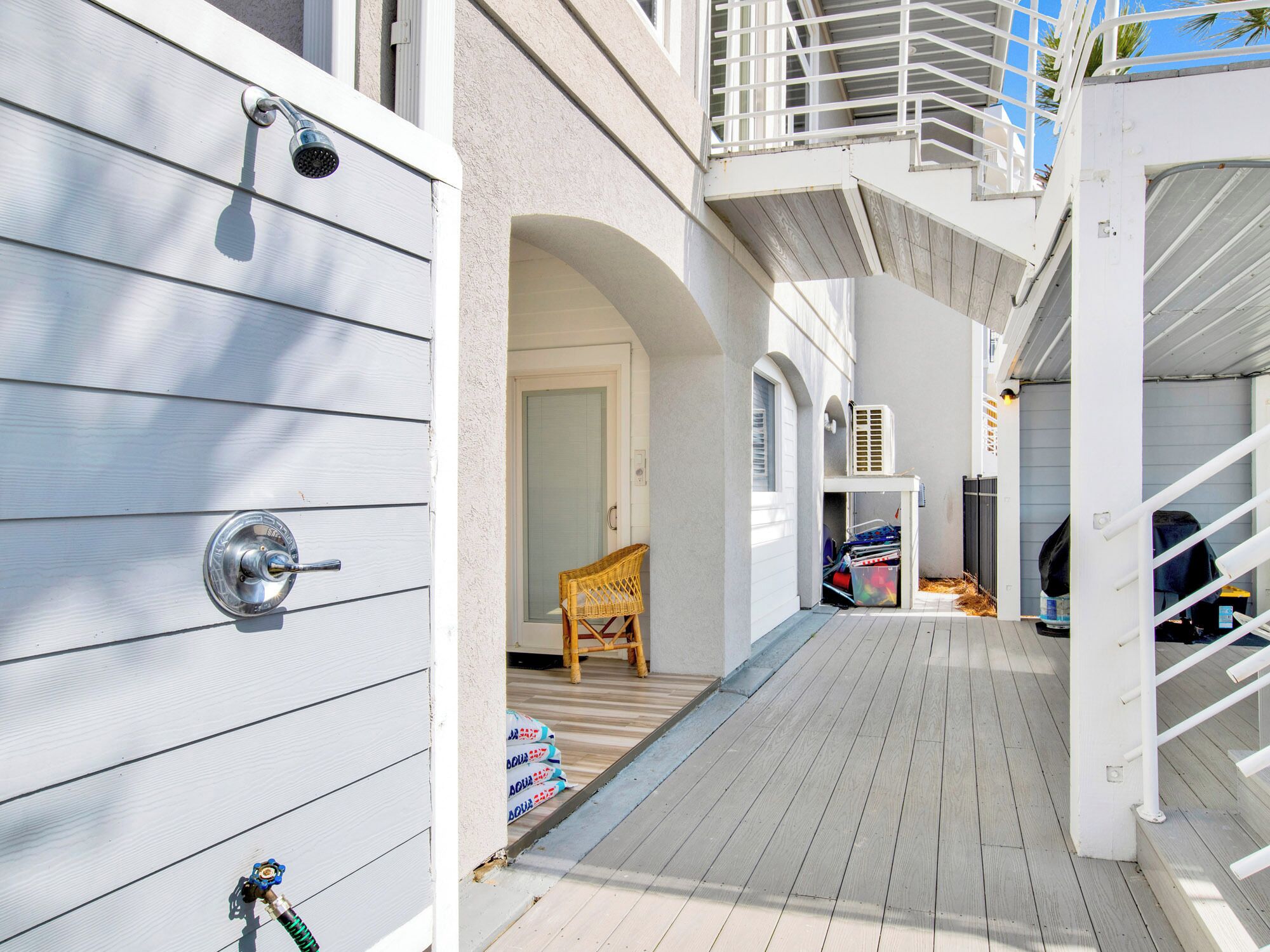 A narrow wooden deck with a white railing, a wicker chair, and a door leading to a covered area with a white air conditioning unit.
