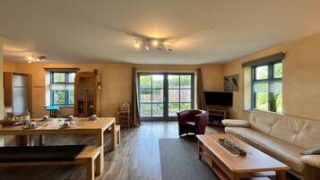 A spacious living room with a sofa, a dining table set for four, a television, and a sliding glass door leading to a garden.