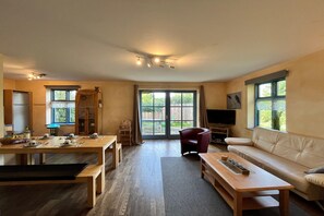 Un salon spacieux comprenant un canapé, une table à manger pour quatre personnes, un téléviseur et une porte-fenêtre coulissante donnant sur un jardin.