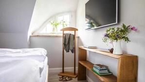 Un dormitorio con una cama, una estantería de madera con libros y un jarrón de flores, un perchero y un televisor montado en la pared.