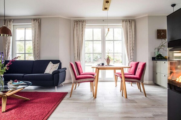A modern living room with a dark sofa, a red rug, a glass coffee table, and a dining area with wooden chairs and a table.