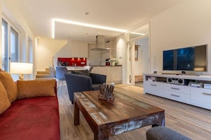 Ein modernes Wohnzimmer mit einer Couch, einem Couchtisch, einem Fernseher und einer Küche im Hintergrund.
