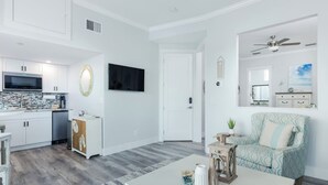 Cocina moderna con armarios blancos, un microondas y un lavavajillas. Hay una zona de comedor con una mesa blanca y un sillón estampado. Un televisor está montado en la pared y se ve un ventilador de techo.