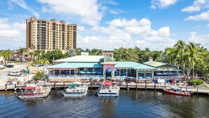 Un restaurante frente al agua con terraza, barcos amarrados y palmeras con un edificio alto al fondo.