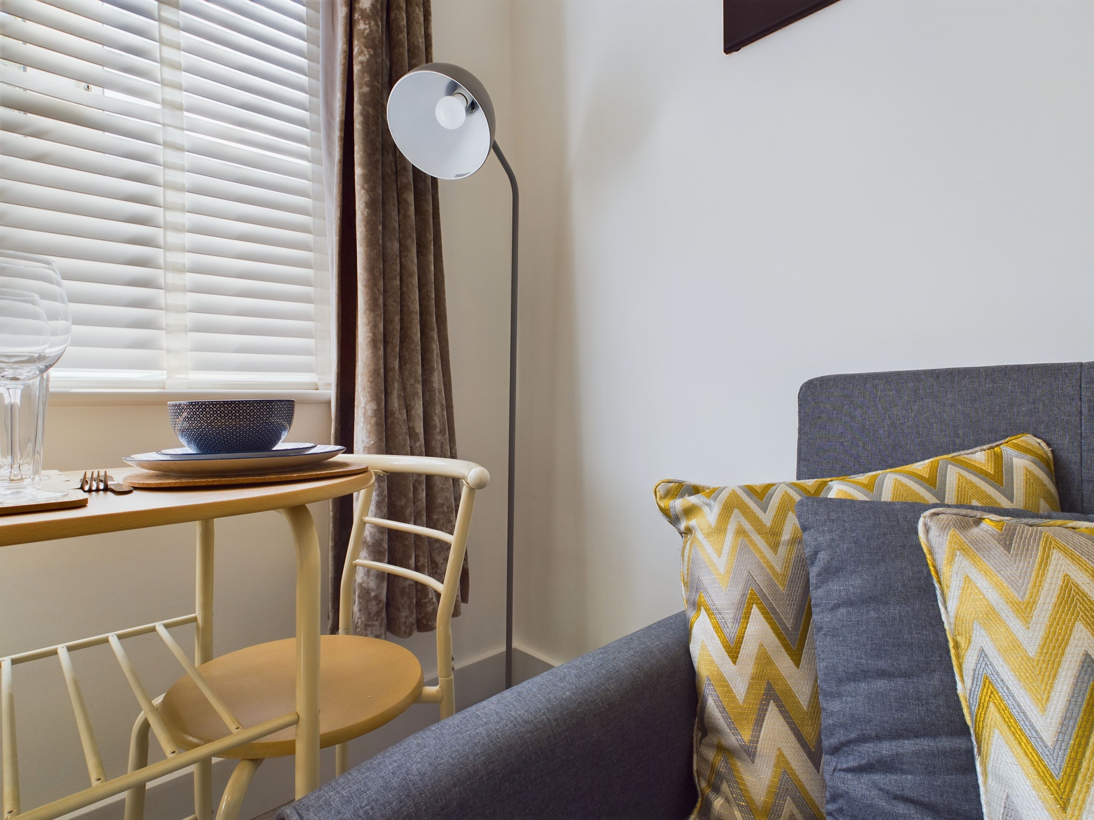 A room with a dining table, a chair, a sofa with patterned cushions, a floor lamp, and a window with blinds.