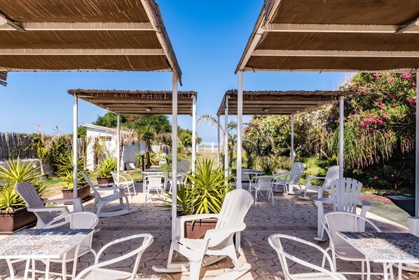 A covered outdoor seating area with white chairs and tables, surrounded by potted plants and a view of the sea.