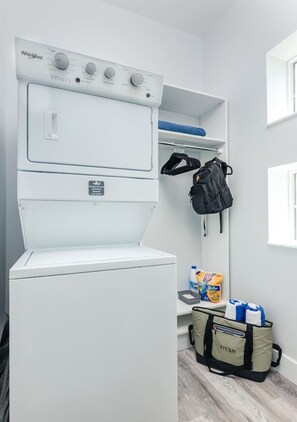 A compact laundry room with a Whirlpool washer and dryer, a shelf with a backpack, and a Titan bag on the floor.