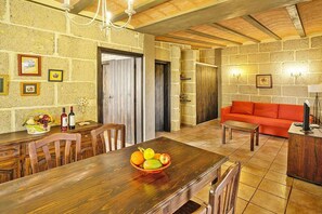 A dining area with a wooden table, chairs, and a fruit bowl. There is a red sofa and a lamp in the living area.