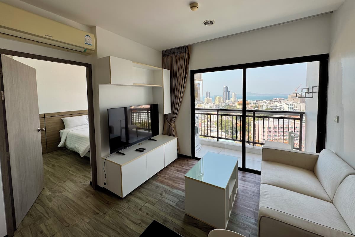 A modern hotel room with a TV on a white cabinet, a sofa, a glass coffee table, and a large window offering a city view.