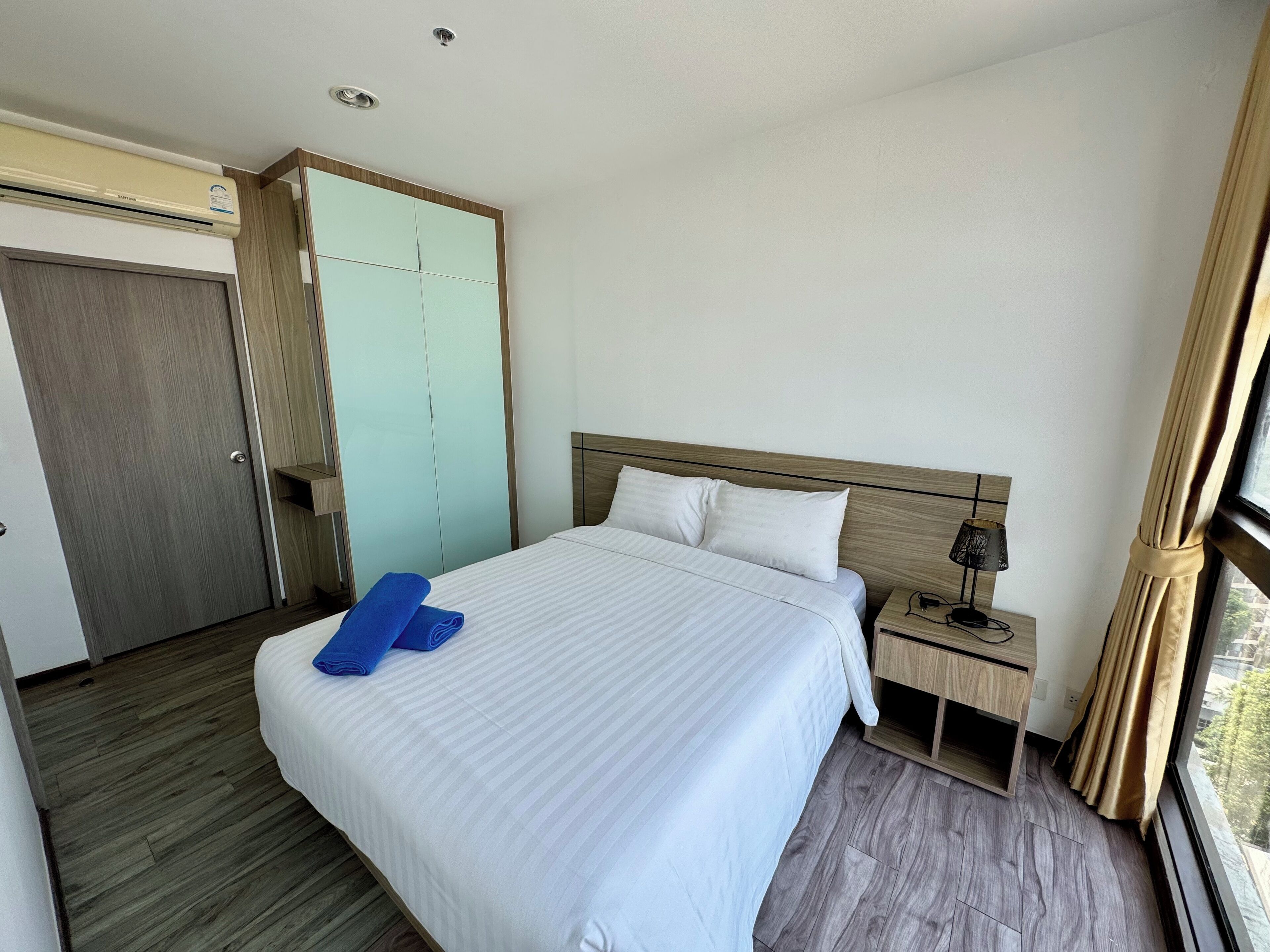 Ein Hotelzimmer mit Bett, Nachttisch, Lampe, Schrank und einem Fenster mit Vorhängen.
