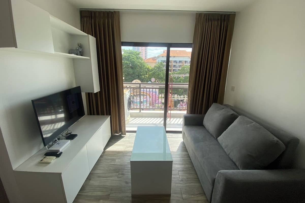 A modern living room with a flat-screen TV, a grey sofa, a white coffee table, and a balcony with a view of buildings and trees.