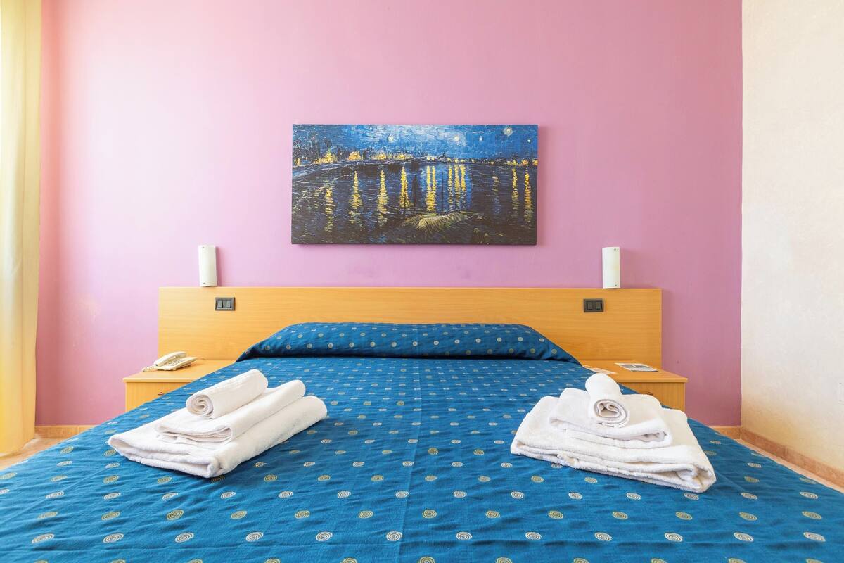 Una habitación de hotel con cama, dos mesitas de noche, un cuadro en la pared y una pared rosa.
