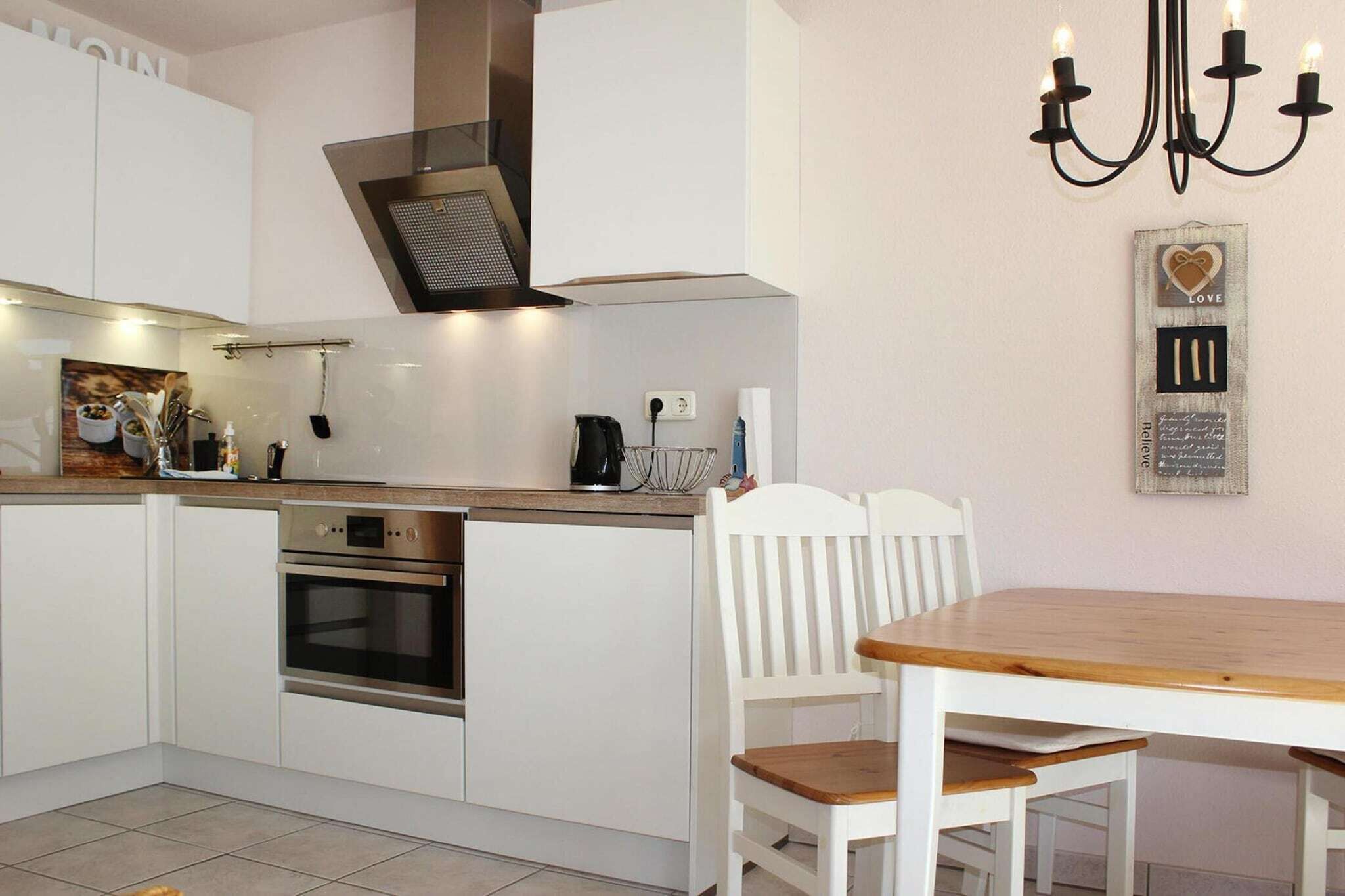 Una cocina moderna con armarios blancos, horno empotrado, mesa de comedor de madera y una araña.