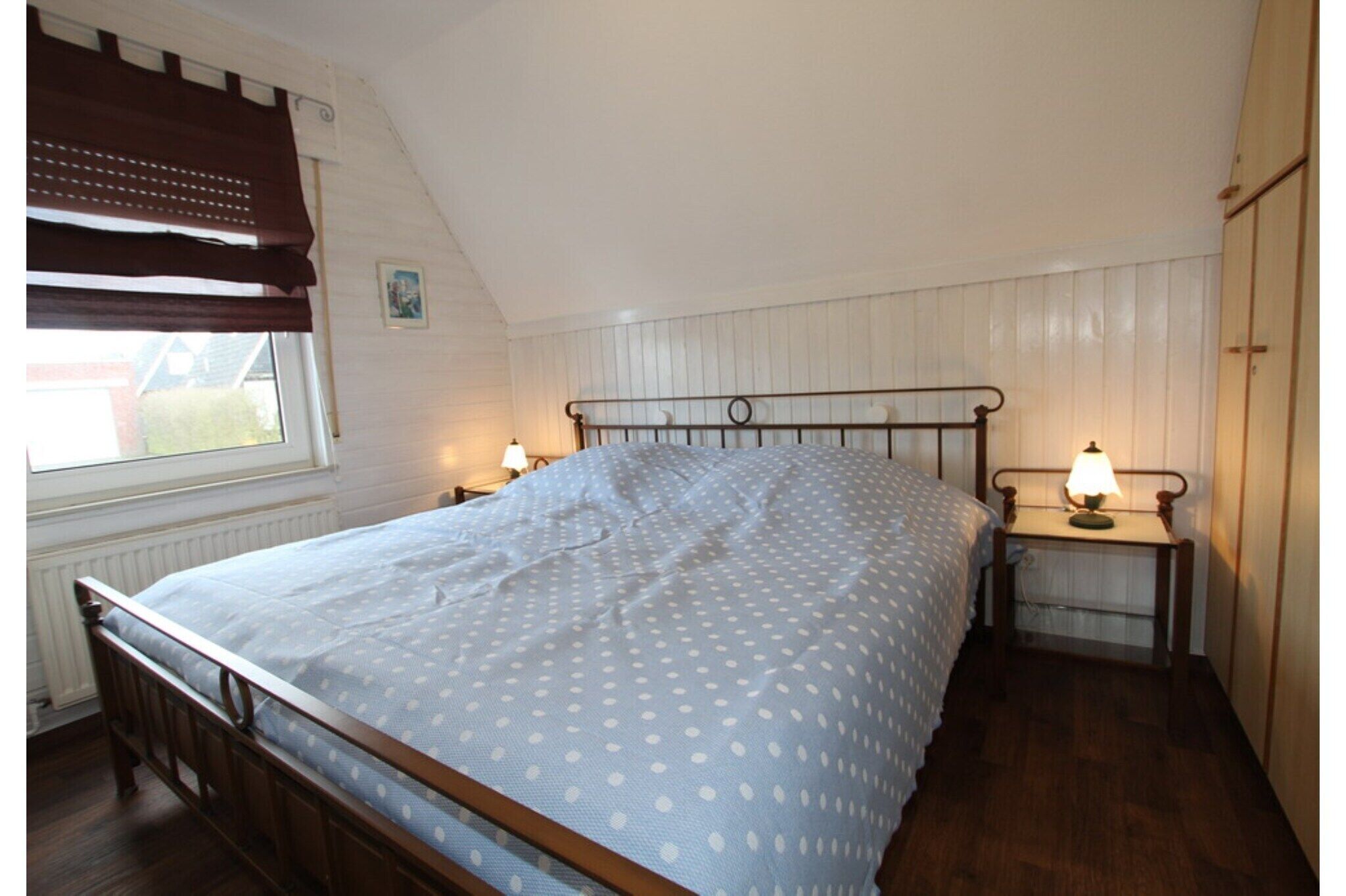 A bedroom with a bed, two bedside tables with lamps, a window with blinds, and a radiator.