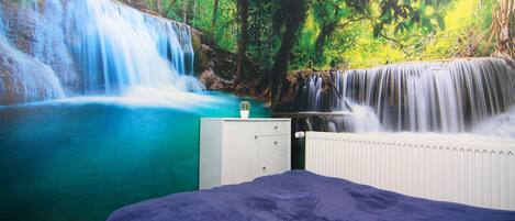Quarto com um grande mural de uma cachoeira e floresta, um penteadeira branca e um edredom azul.