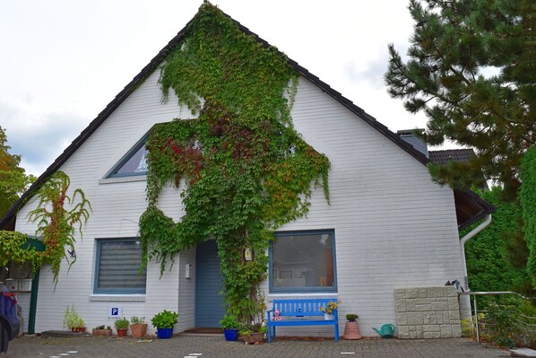 Un edificio blanco con puerta y ventanas azules, cubierto de hiedra verde y un banco con una maceta en la parte delantera.