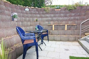 Un patio con una mesa, sillas, un estante pequeño y un muro de piedra con plantas.