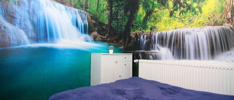 Quarto com um grande mural de uma cachoeira e floresta, um penteadeira branca e um edredom azul.