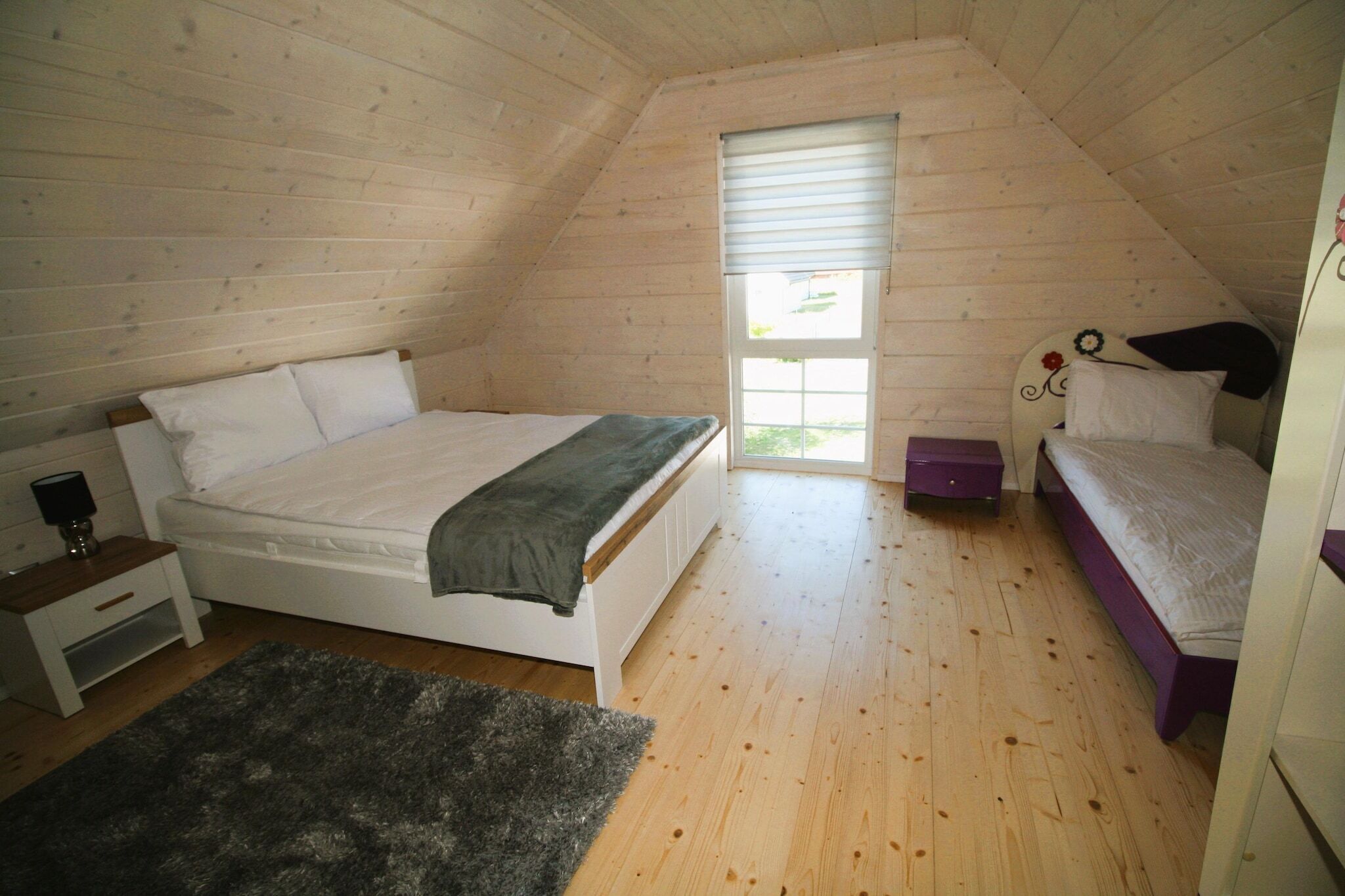 Ein Schlafzimmer mit schrägem Dach, einem Bett mit weißem Kopfteil, einem Nachttisch, einem Fenster mit Jalousien und einer violetten Bank.