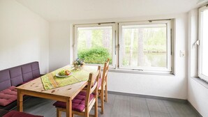 Ein Essbereich mit Tisch für zwei Personen, Holzsesseln, einer Bank mit Kissen und großen Fenstern mit Blick ins Grüne.