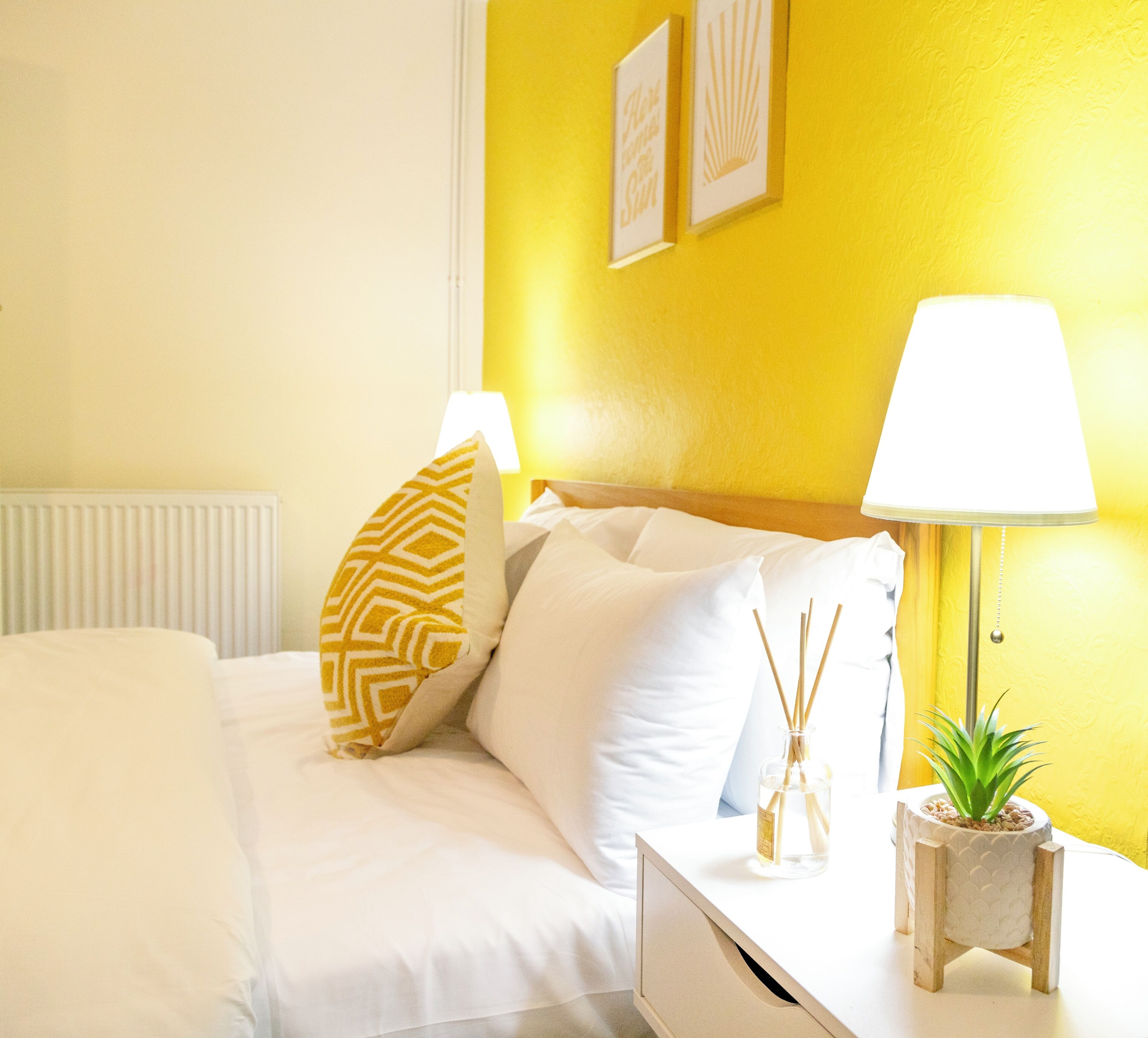 A neatly made bed with white bedding and patterned pillows, a bedside table with a lamp and a small plant, and a yellow wall with two framed pictures.
