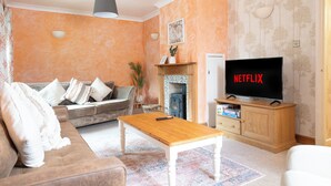 A living room with a sofa, a coffee table, a TV displaying 'NETFLIX', and a fireplace. - Charming Frome Retreat With 4 Bedrooms (Frome)