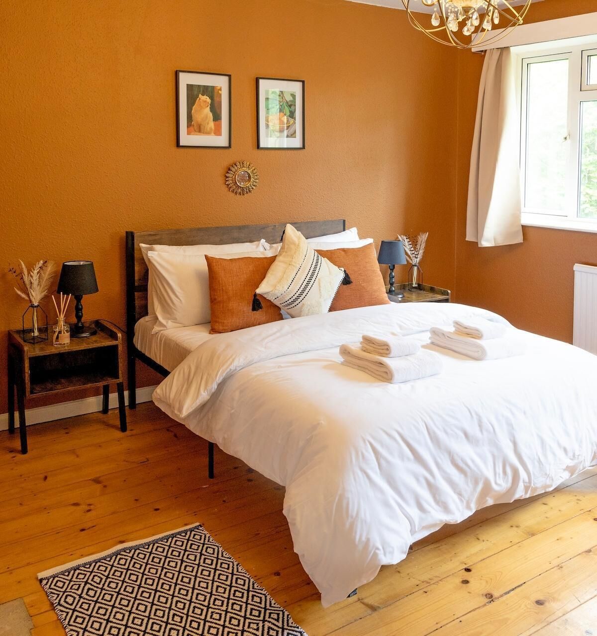A neatly made bed with white linens and towels, a wooden headboard, a patterned rug, and two framed pictures on the wall.