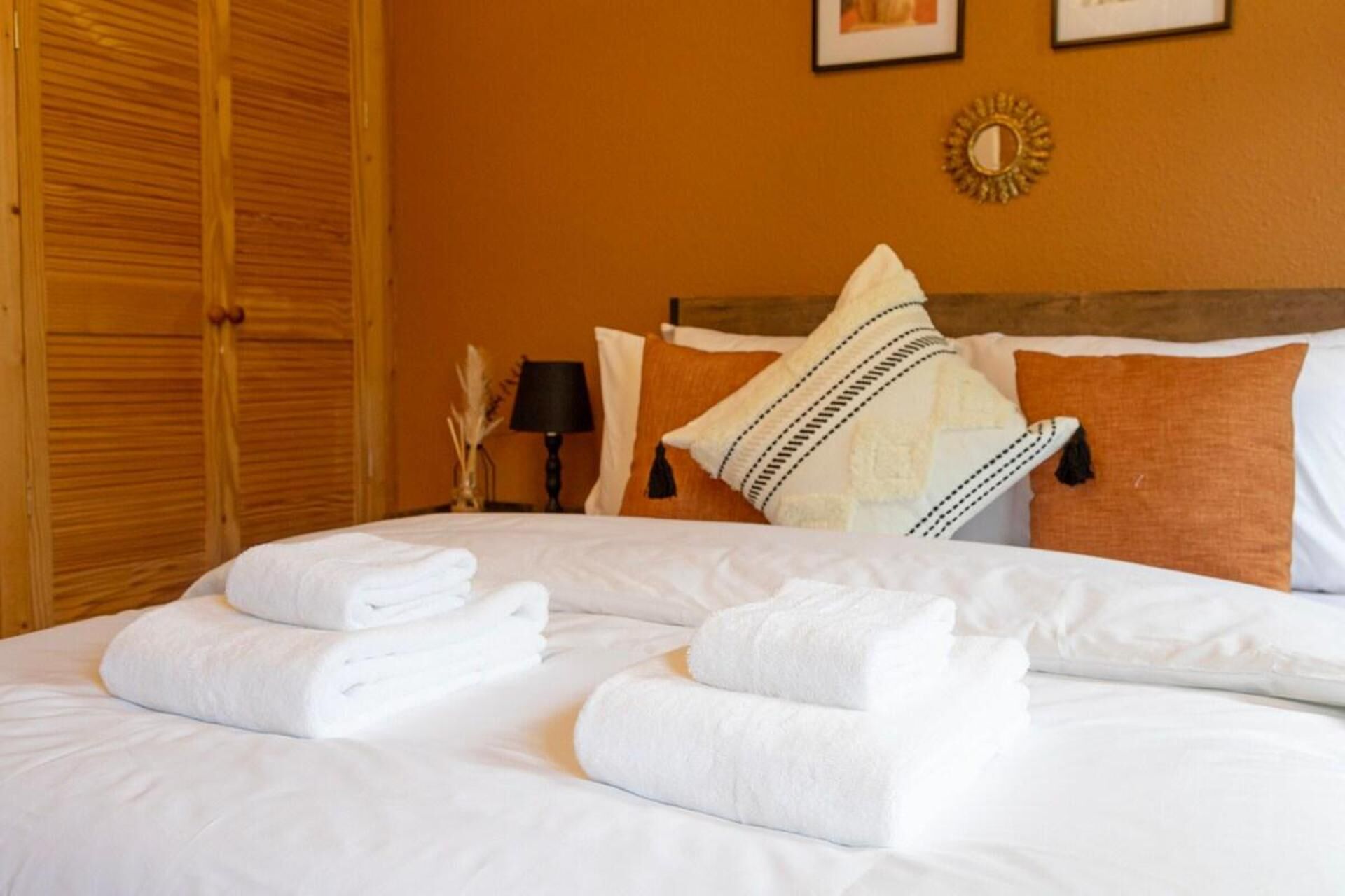 A neatly made bed with white linens and towels, a wooden wardrobe, and a wall-mounted mirror.