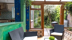 A patio area with wicker furniture, a small table with a plant, and a view of a garden with a barbecue grill. - Charming Frome Retreat With 4 Bedrooms (Frome)