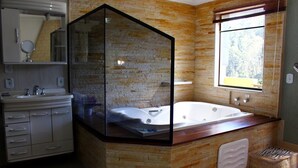 A bathroom with a stone wall, a glass-enclosed shower, a white bathtub, and a window with a view of trees. - POUSADA BELA VISTA (Camanducaia)