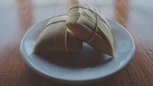 A plate with two tamales on a wooden table, with a window in the background. - POUSADA BELA VISTA (Camanducaia)