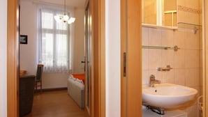 Une chambre d’hôtel avec un plancher en bois, un lit blanc, une chaise, une petite table, une fenêtre avec des rideaux légers et une salle de bain avec un lavabo et un miroir blancs.